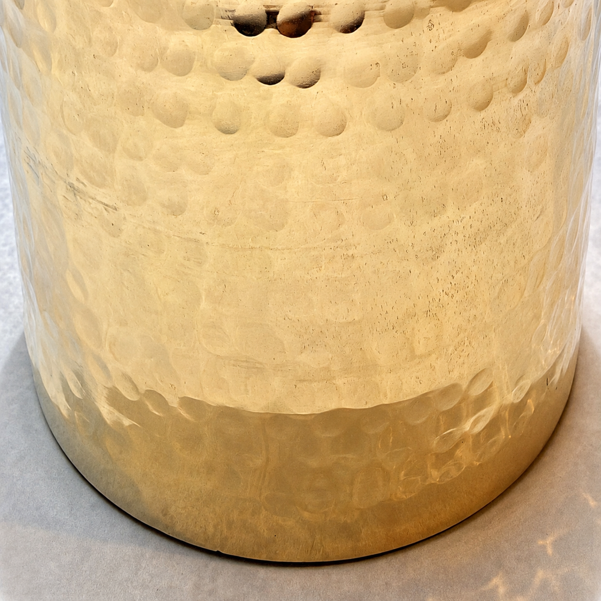 Close-up of a hammered metal cylindrical container or vessel base in a warm metallic gold finish, showing the textured surface and dimpled pattern.