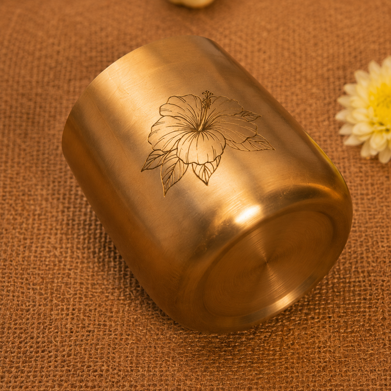 A brushed brass cylindrical cup or tumbler with an engraved hibiscus flower design, resting on a brown burlap fabric background with a small white flower prop in the corner.