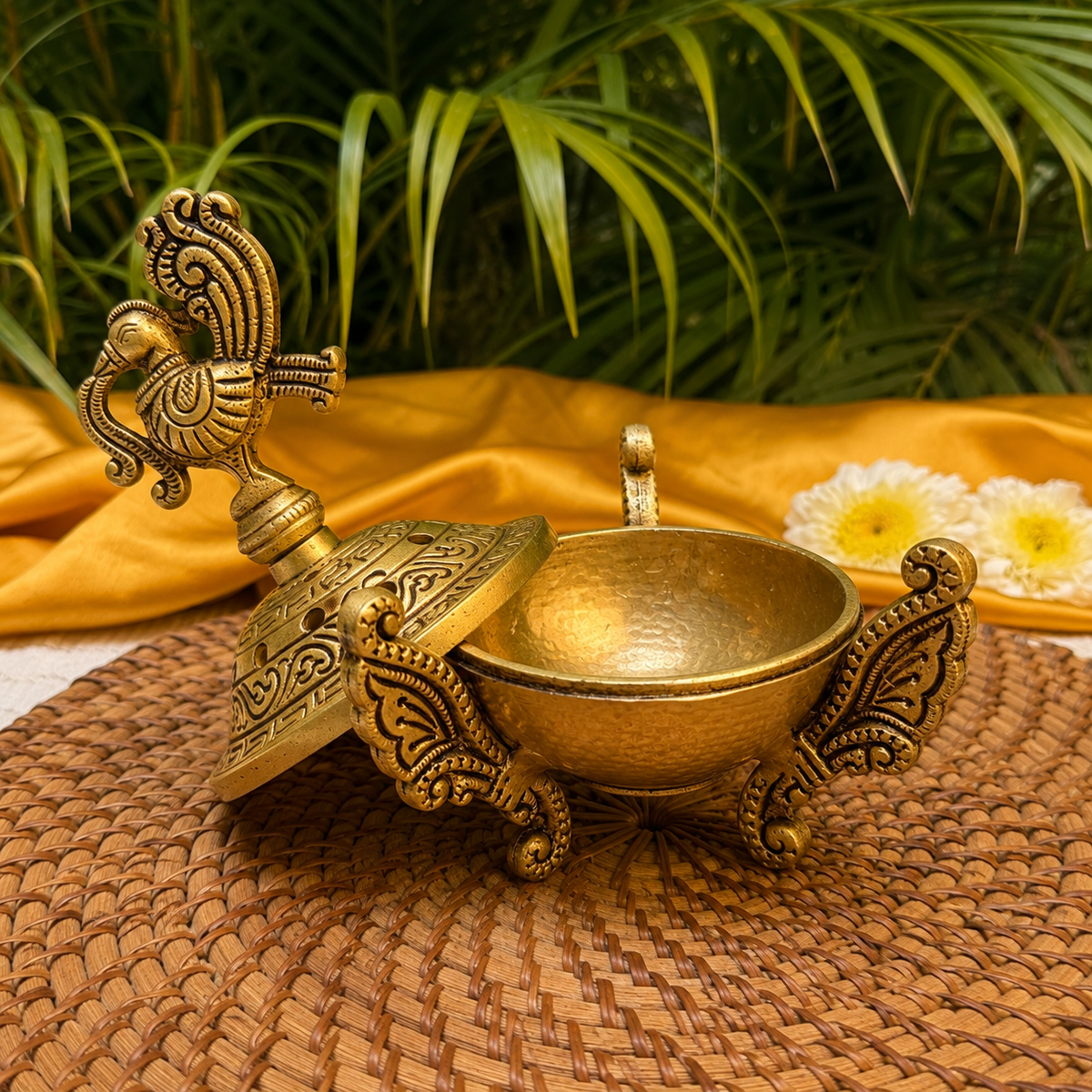 An ornate brass oil lamp with a matching decorative lid, styled on a woven mat with a draped yellow fabric and green leaves in the background.