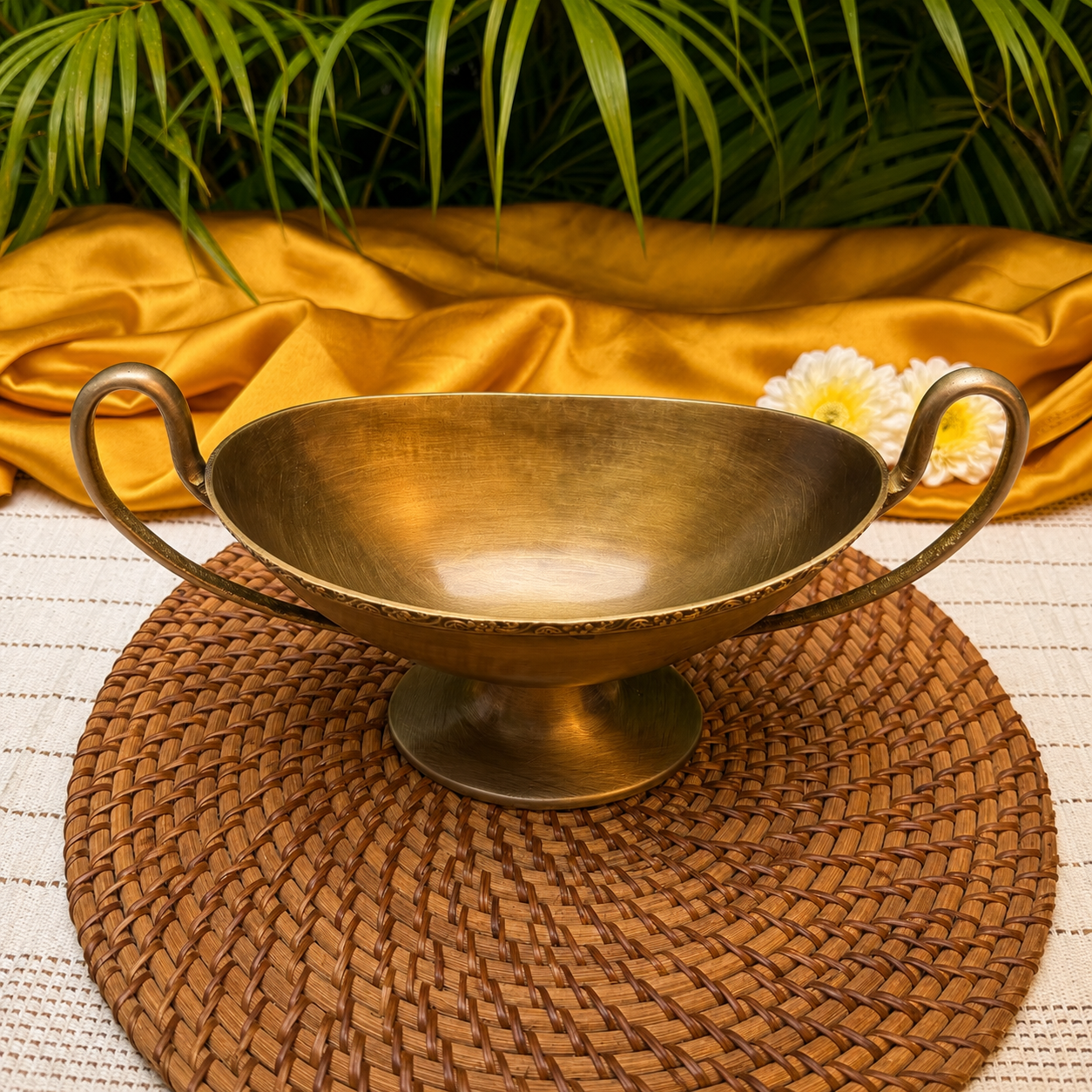 A brass-colored double-handled bowl with a pedestal base, styled on a woven brown placemat with gold satin fabric, green palm leaves, and a white flower in the background.