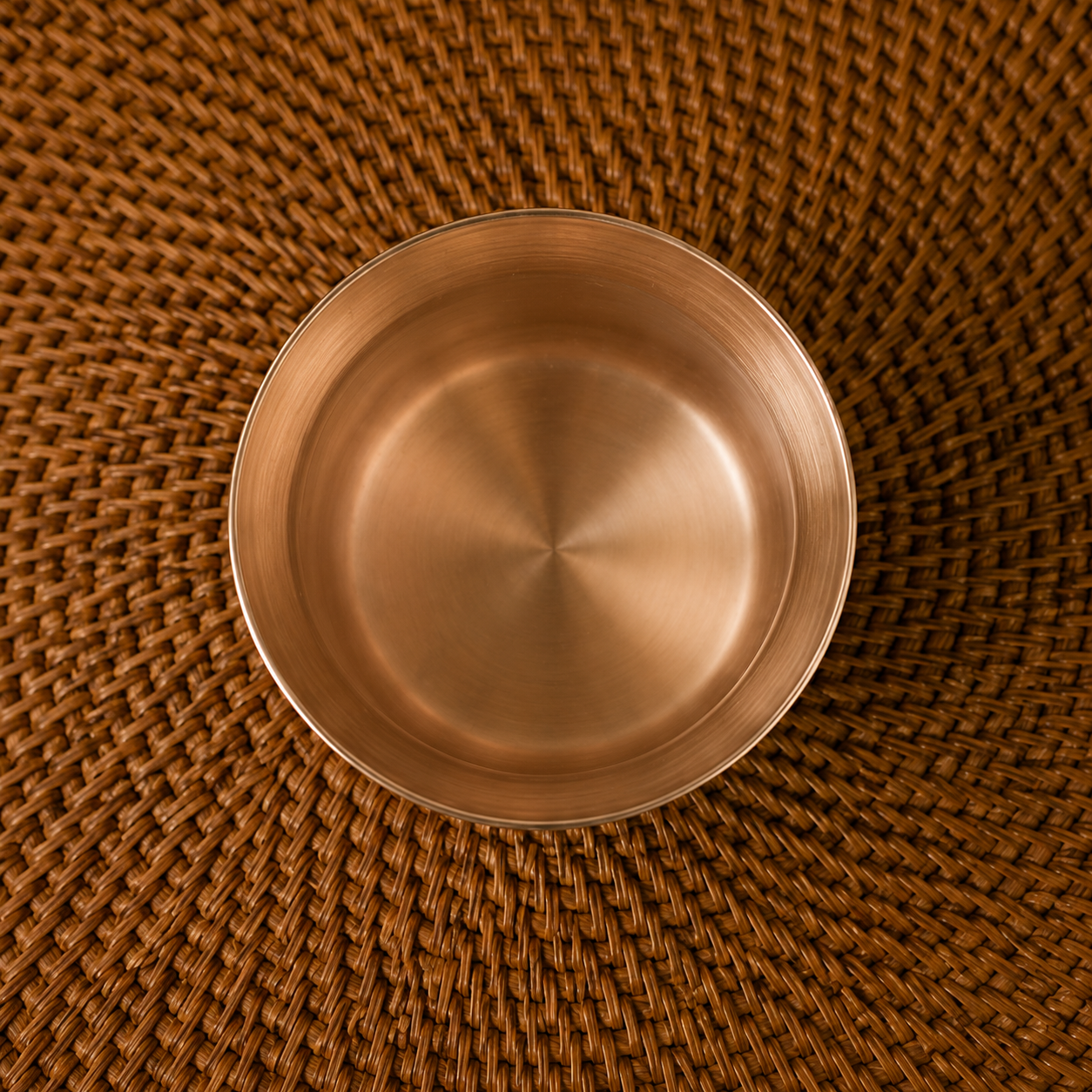 A round brushed metal bowl with a warm copper or bronze finish, photographed from directly above on a brown woven placemat.