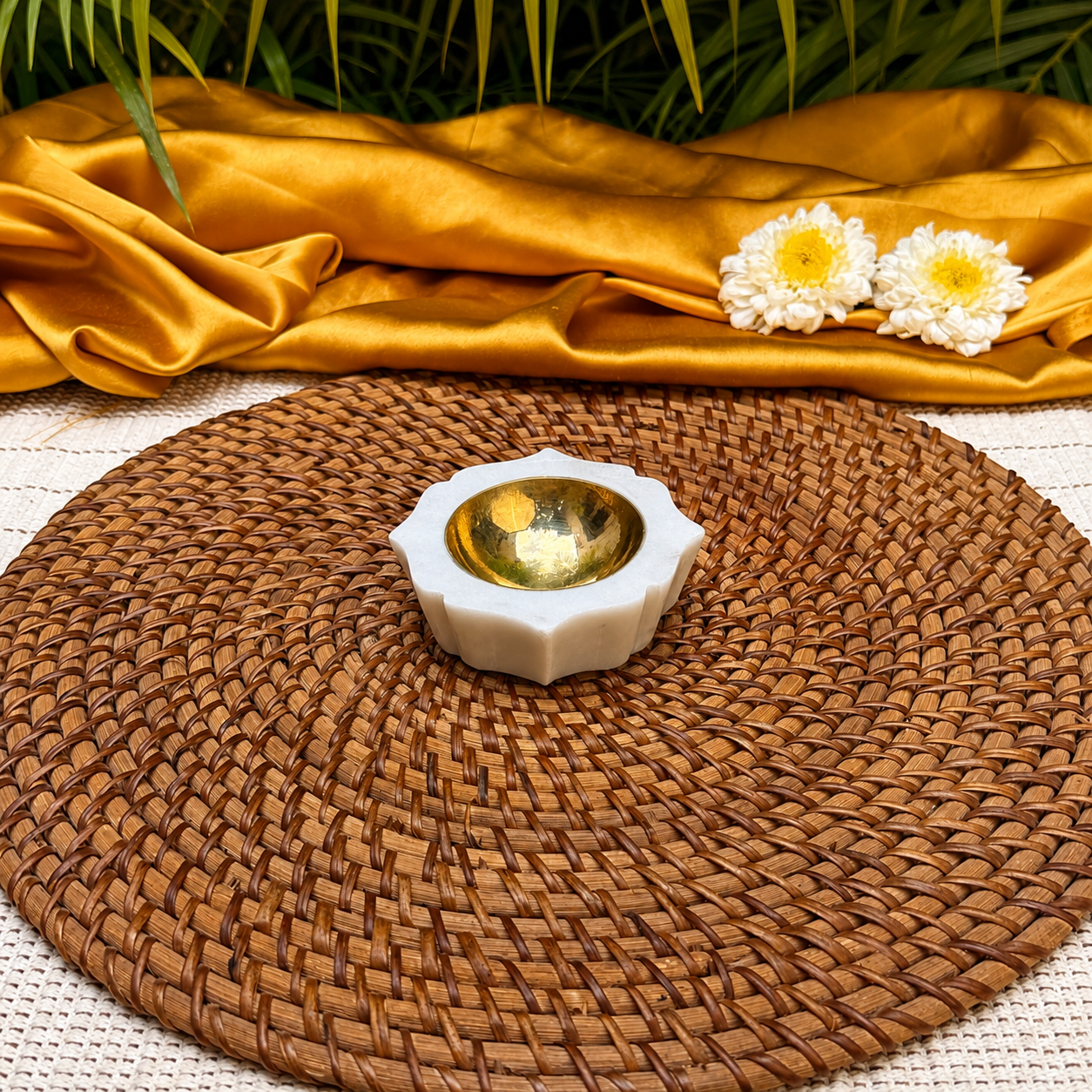 A small white ceramic candle holder with a reflective gold interior, placed on a round woven brown placemat. Gold satin fabric and two white daisy flowers are draped behind it, with green leaves visible at the top edge.