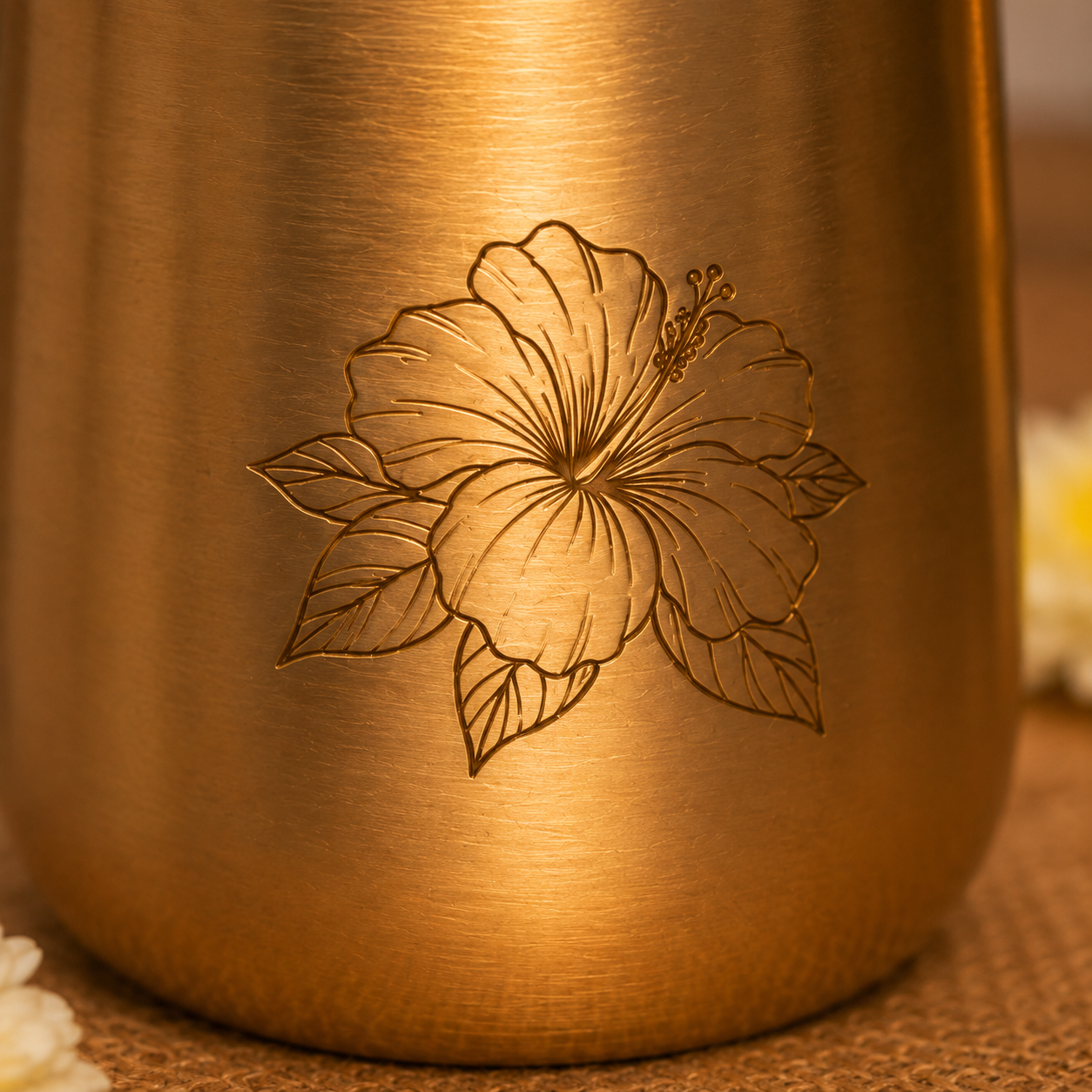 Close-up of a brushed metallic gold tumbler with an engraved hibiscus flower design, resting on a textured surface with soft blurred flowers in the background.