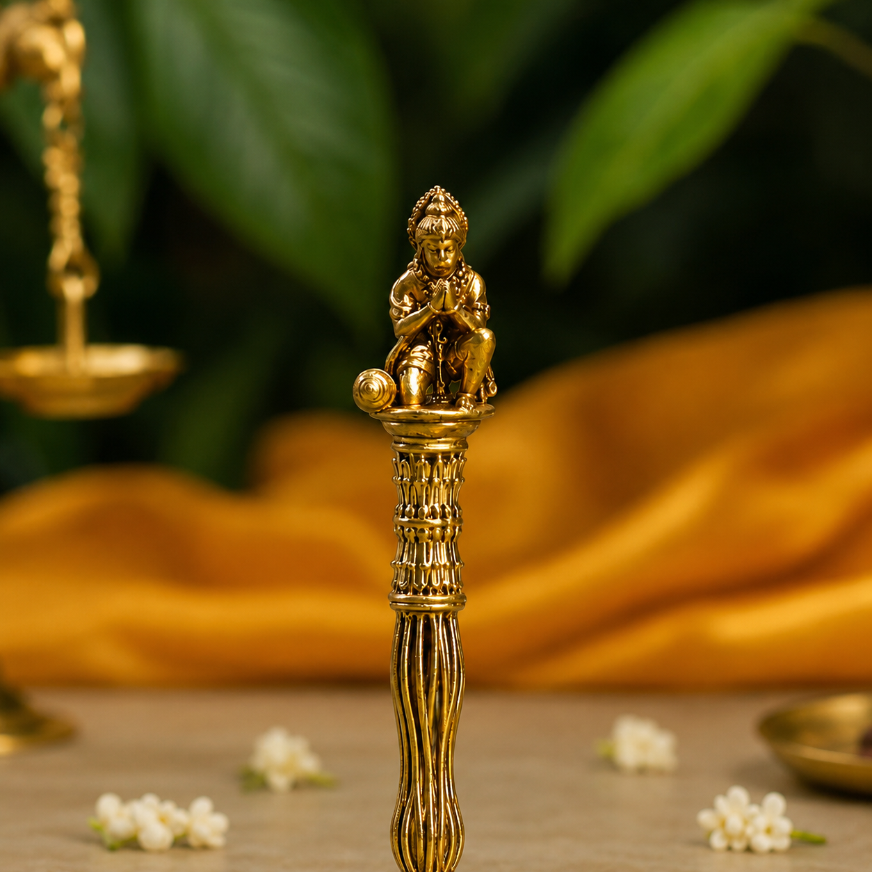A brass-toned decorative object with a small seated figure sculpture on top, standing on a textured pedestal. The item is photographed on a tabletop with blurred green leaves and draped golden fabric in the background.