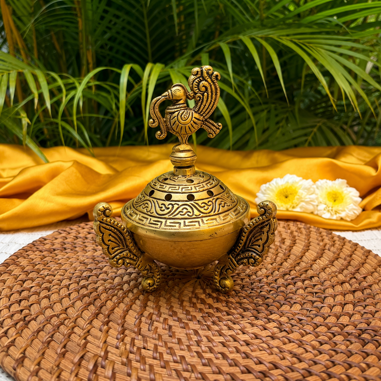 An ornate brass incense burner with a decorative peacock finial on top, set on a woven brown mat with draped yellow fabric and green palm leaves in the background.