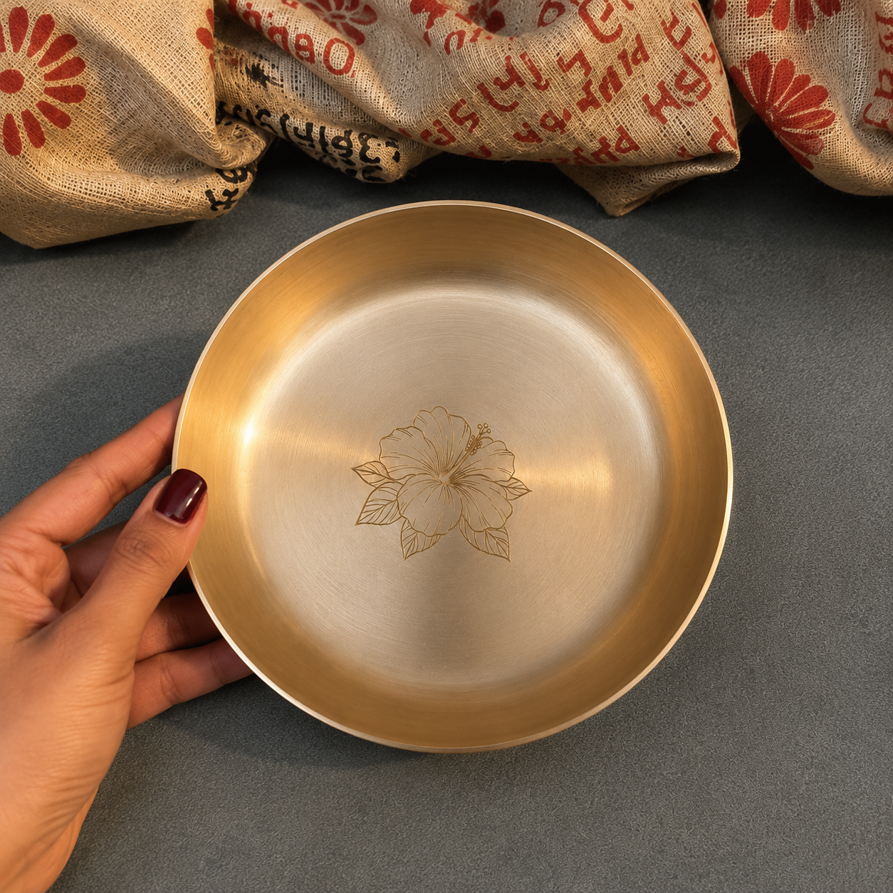 A hand with dark red nail polish holds a round, brushed gold metal bowl or plate with an engraved floral design in the center. The background is a textured gray surface with decorative burlap fabric draped at the top.