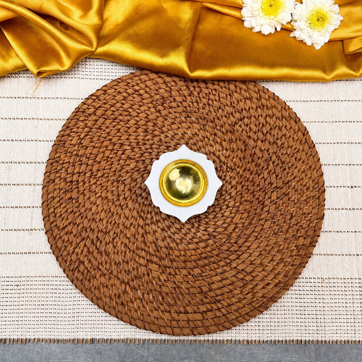 A round brown woven placemat or charger plate centered on a white textured table runner, with a small white and gold dish in the middle. Gold satin fabric and two white daisy flowers are draped across the top edge.