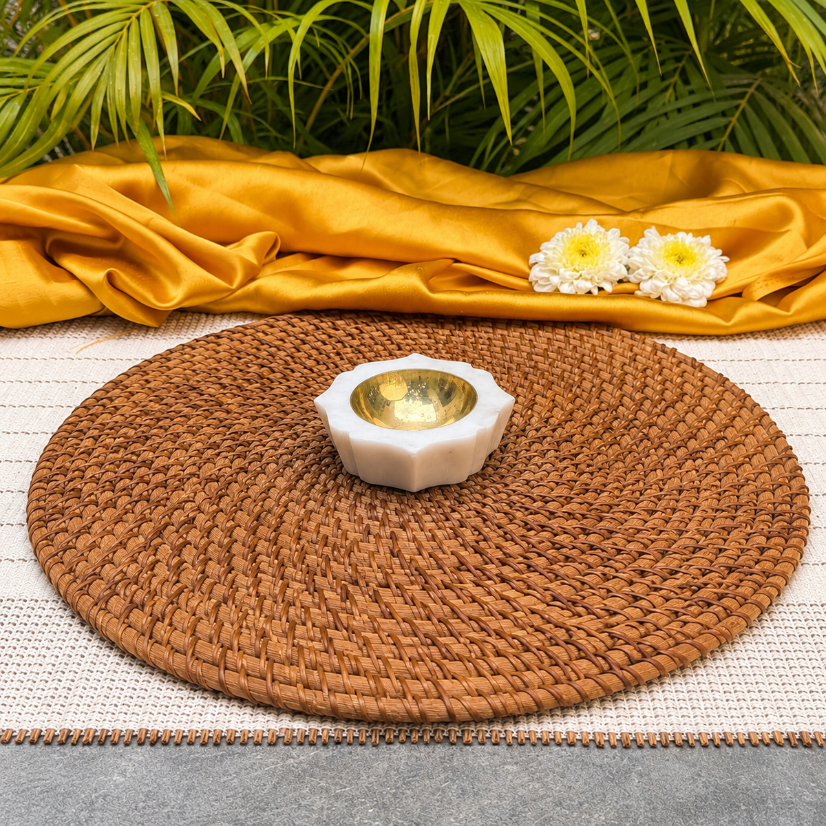 A round brown woven placemat on a white woven runner, styled with a small white and gold candle holder, white flowers, and a draped golden silk cloth with green leaves in the background.