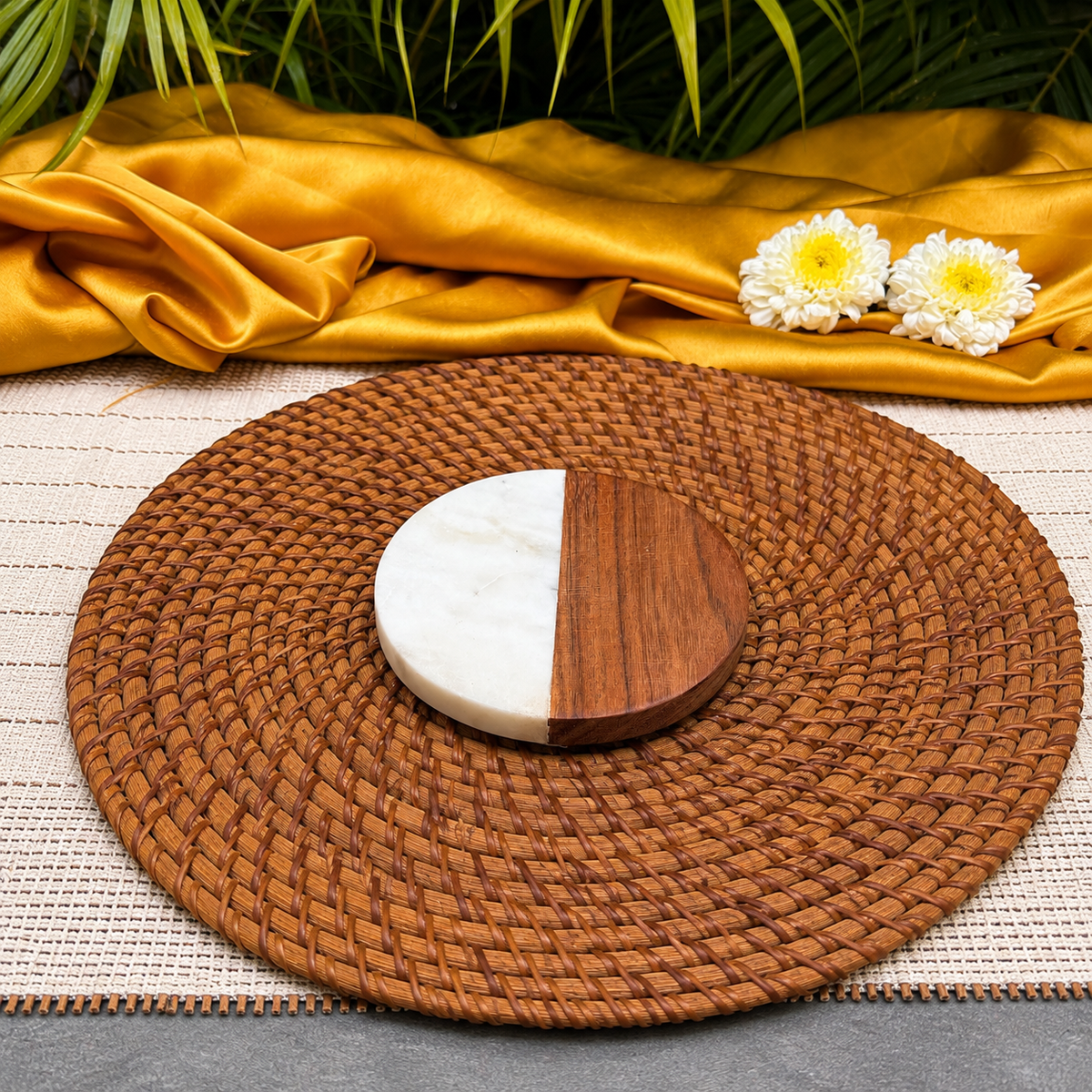 A round woven brown placemat on a light woven mat, with a round wooden coaster or tray in half white marble and half medium brown wood, styled with a gold satin cloth, green leaves, and two white flowers.
