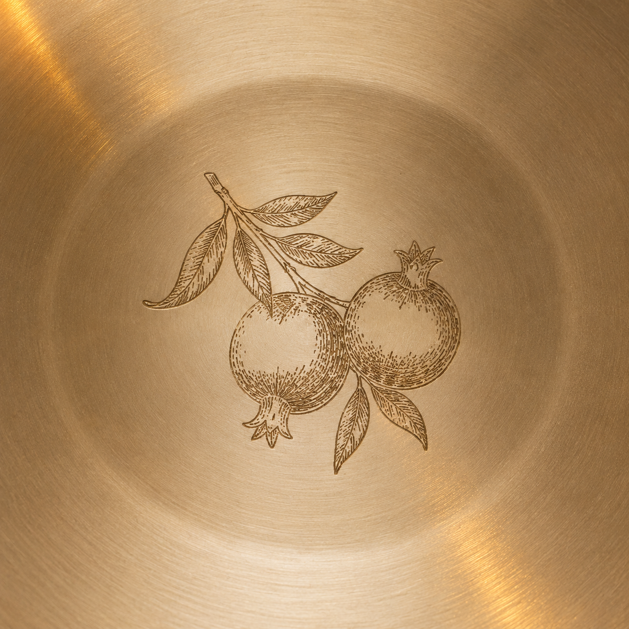 Close-up of a brushed metallic surface with an engraved illustration of two pomegranates on a branch, showing warm gold and bronze tones.