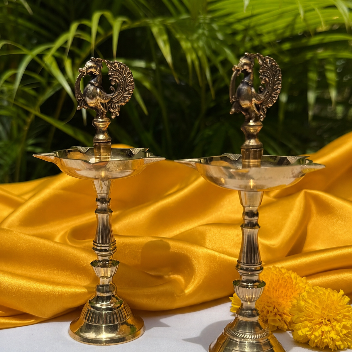 Two ornate brass oil lamps with peacock figurines on top, displayed on draped yellow satin fabric with green foliage in the background.