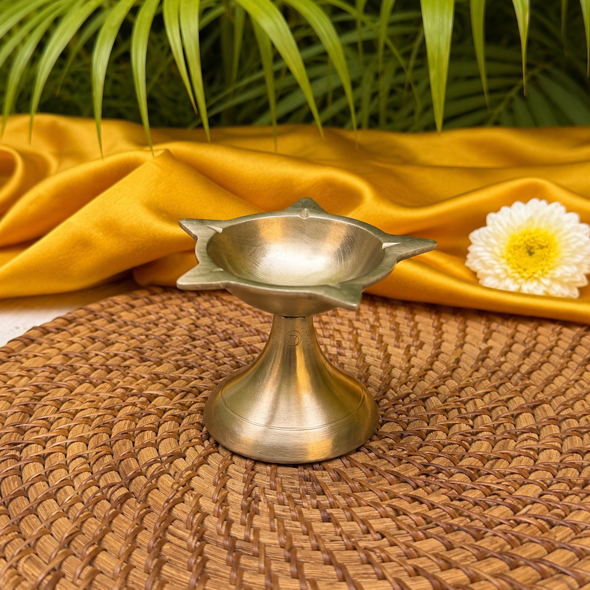 A brass diya holder with a star-shaped rim and pedestal base, styled on a woven brown mat with a draped golden-yellow fabric, green leaves, and a white flower in the background.