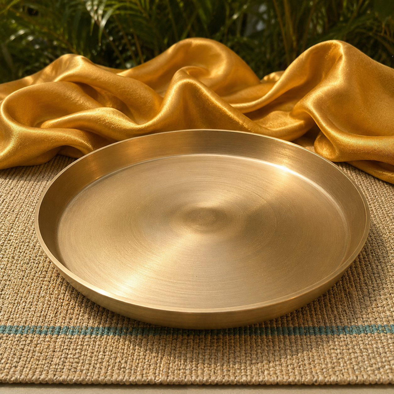 A round brushed brass serving platter or shallow tray, styled on a woven beige mat with draped gold satin fabric and green foliage in the background.