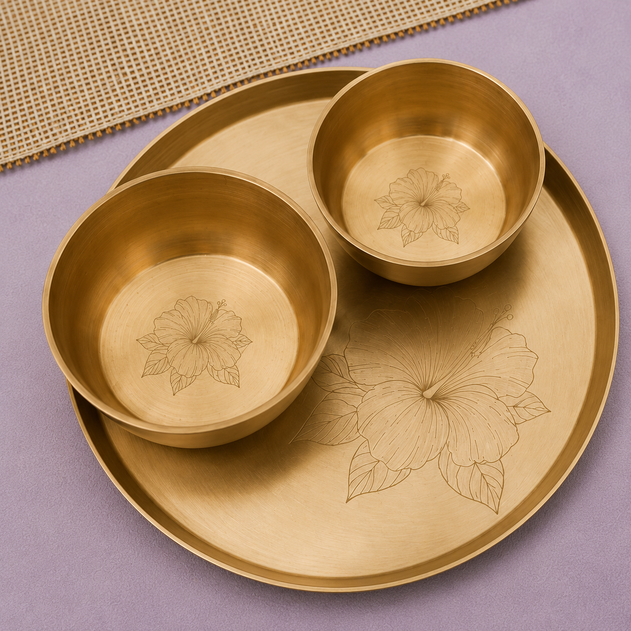A brass serving tray with two matching small bowls, both engraved with hibiscus flowers, displayed on a lavender surface with a woven mat in the background.