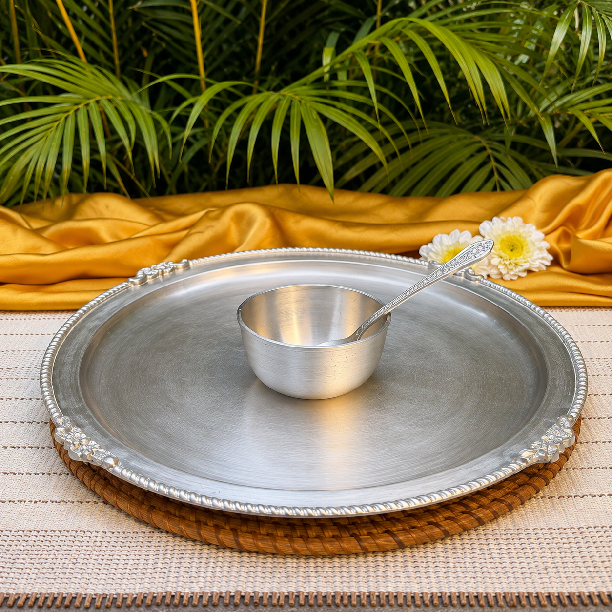 A silver metal serving tray with decorative corner details and a matching small bowl and spoon, arranged on a woven placemat with gold fabric and green palm leaves in the background.