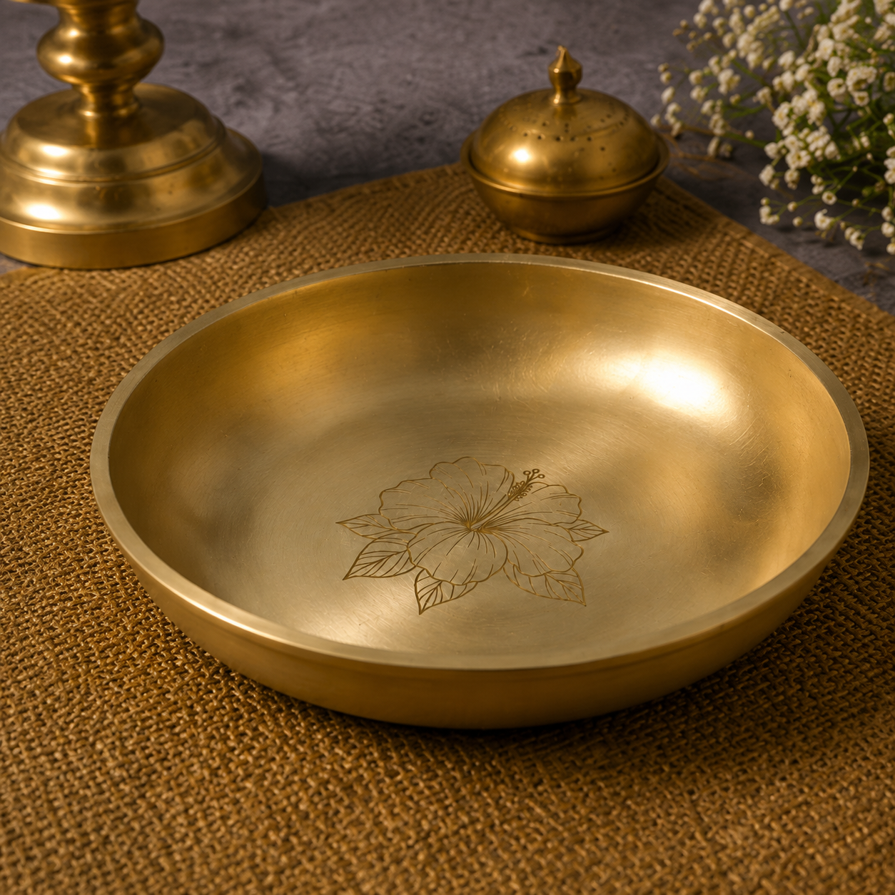 A round brass bowl with a hand-engraved hibiscus flower design inside, displayed on a textured brown woven mat with decorative brass and floral props in the background.