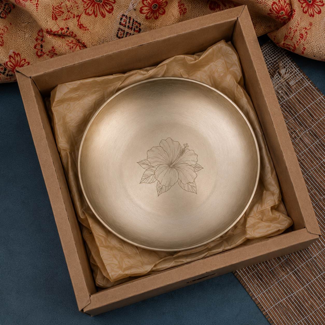 A round brushed-metal plate or tray with an engraved floral motif in the center, presented inside an open brown cardboard box lined with tissue paper.