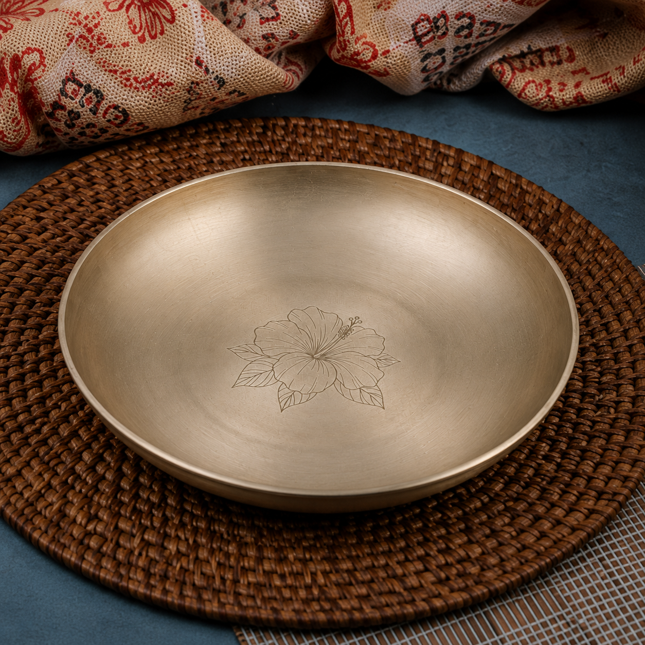 A round brushed-metal bowl with an engraved hibiscus flower in the center, resting on a brown woven placemat over a blue surface with patterned fabric in the background.
