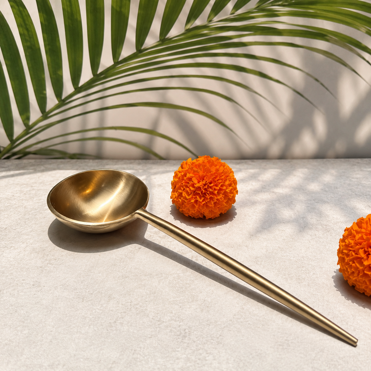 A long-handled brass spoon with a polished golden finish, styled on a light textured surface with orange marigold flowers and a green palm leaf shadow in the background.