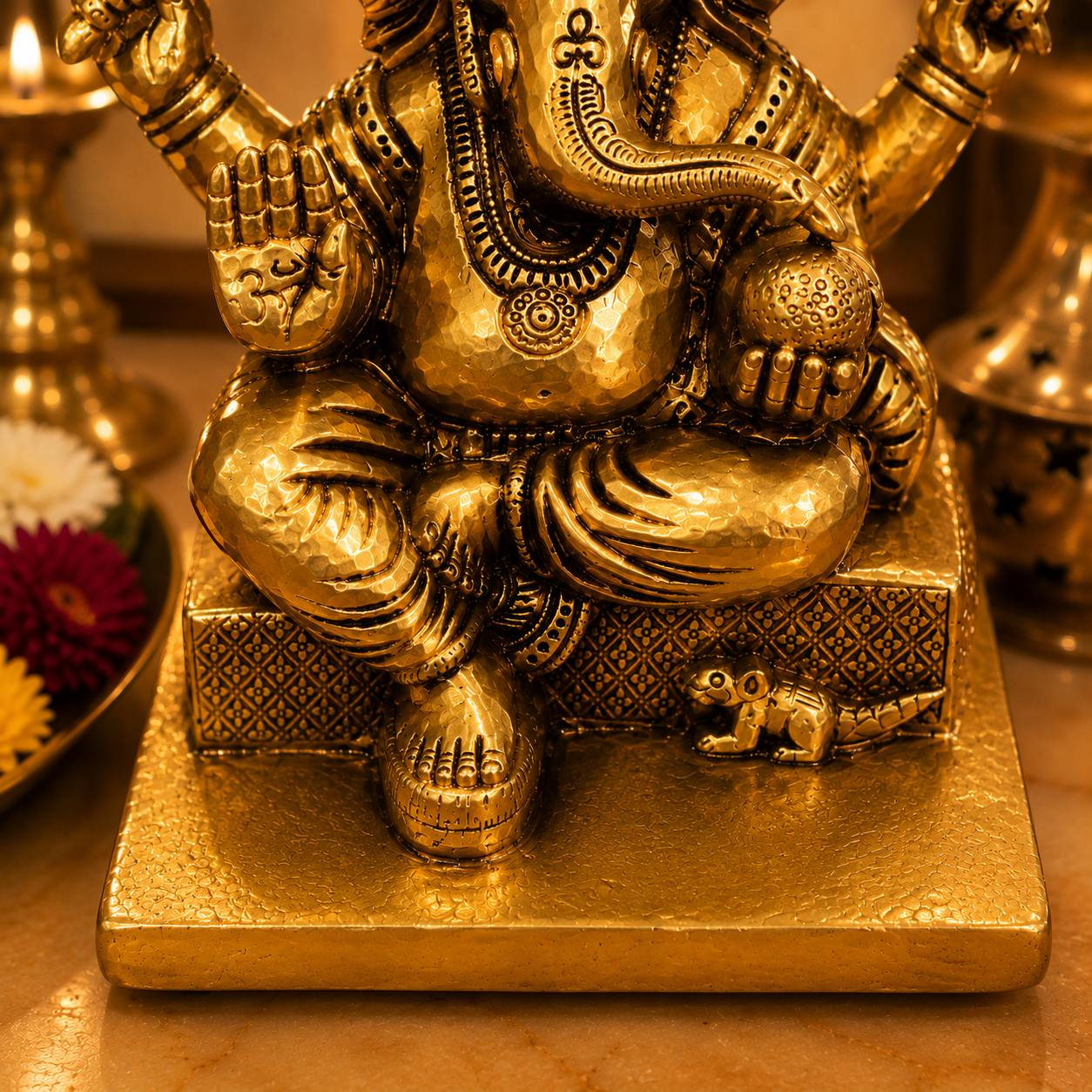 A close-up of a gold-toned Ganesha statue seated on a pedestal, with ornate detailing and a small monkey figure beside it, photographed indoors with warm ambient lighting.
