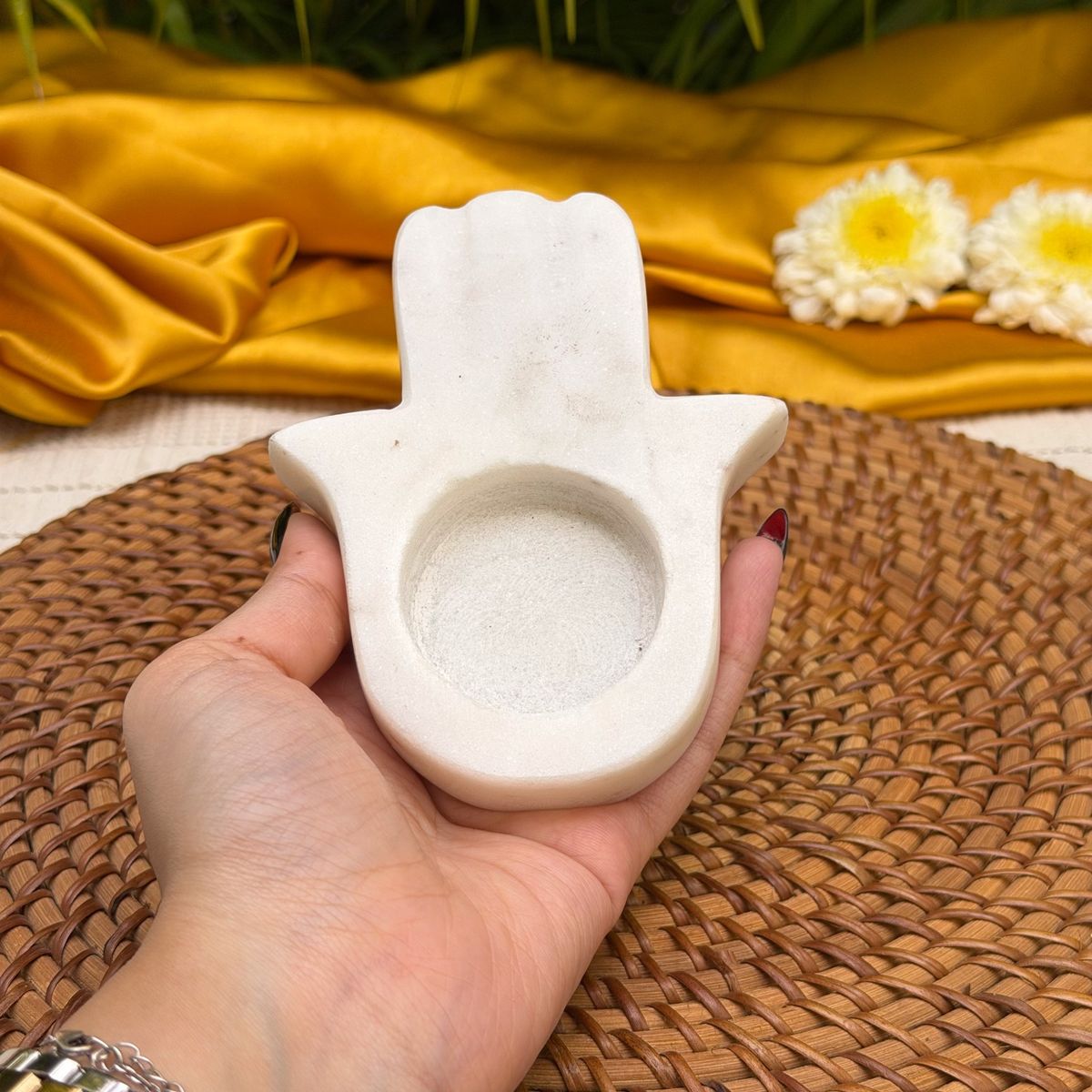A hand holds a small white stone hamsa hand container filled with white powder, set against a woven brown placemat with a gold satin cloth and white flowers in the background.