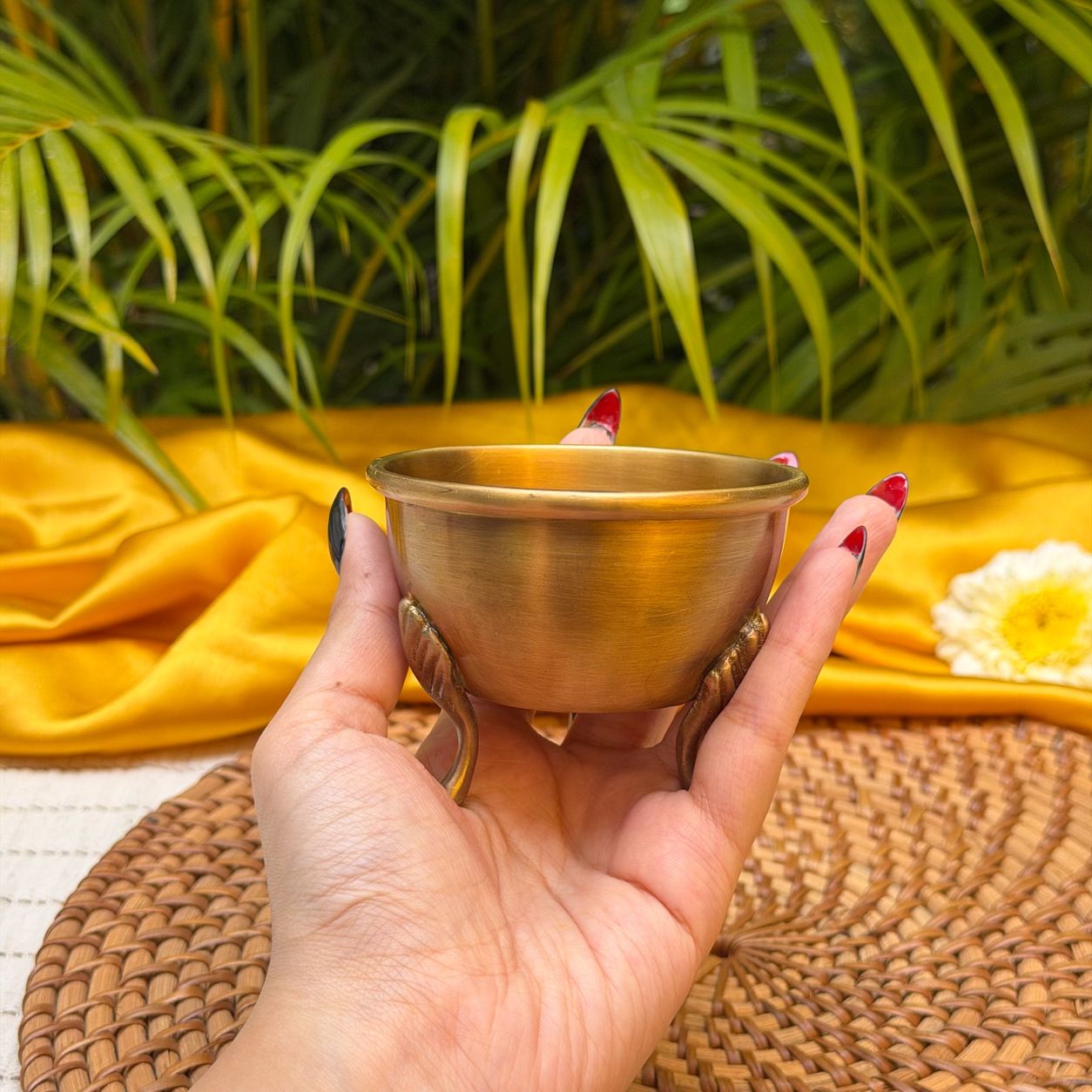 A brass cup held in a person's hand, with green palm leaves and a yellow fabric backdrop in the background.