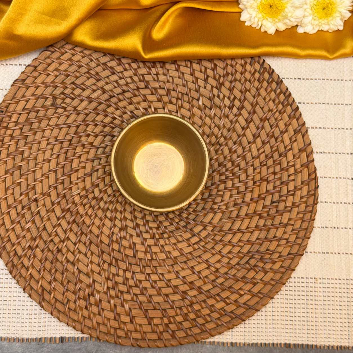 A round woven brown placemat with a small metallic gold bowl in the center, styled on a cream woven mat with gold fabric and white flowers at the top.