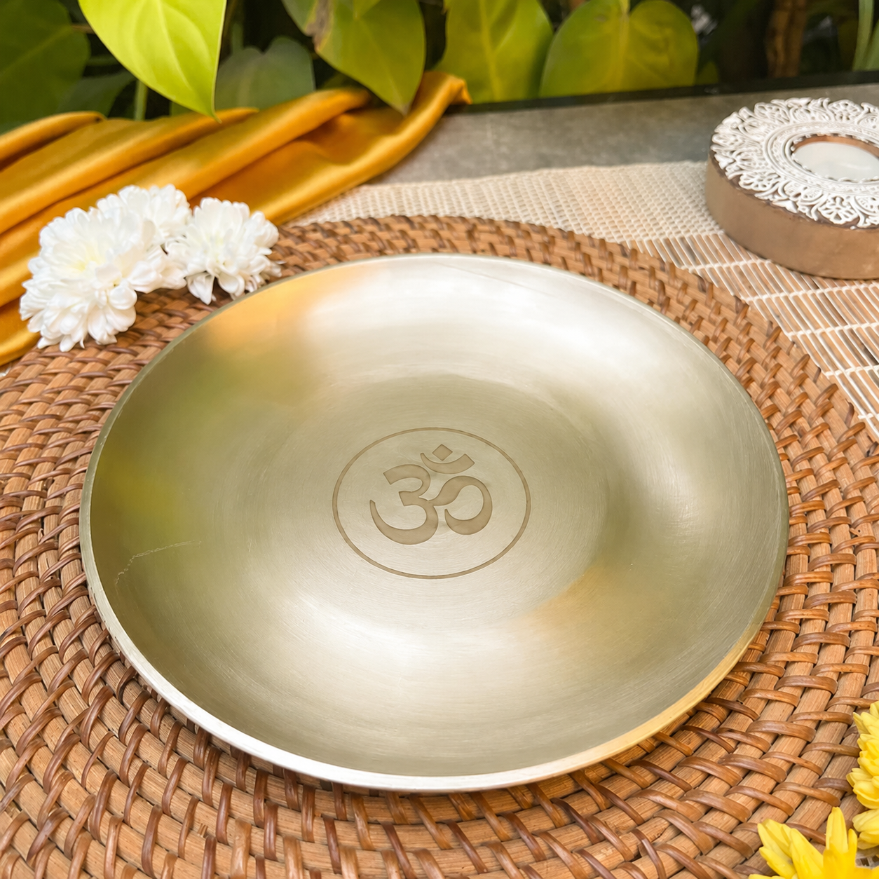 A round metallic brass plate with an engraved Om symbol in the center, displayed on a woven brown placemat with decorative white flowers and green leaves in the background.