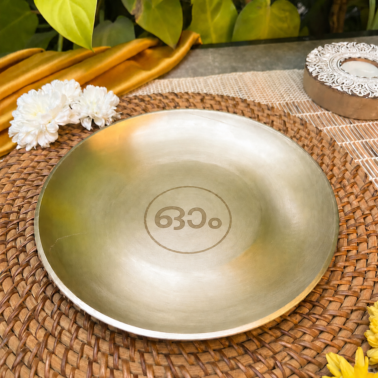 A round metallic plate with a brushed gold finish and the engraved text 'om.' in the center, resting on a woven brown placemat with decorative flowers and green leaves in the background.