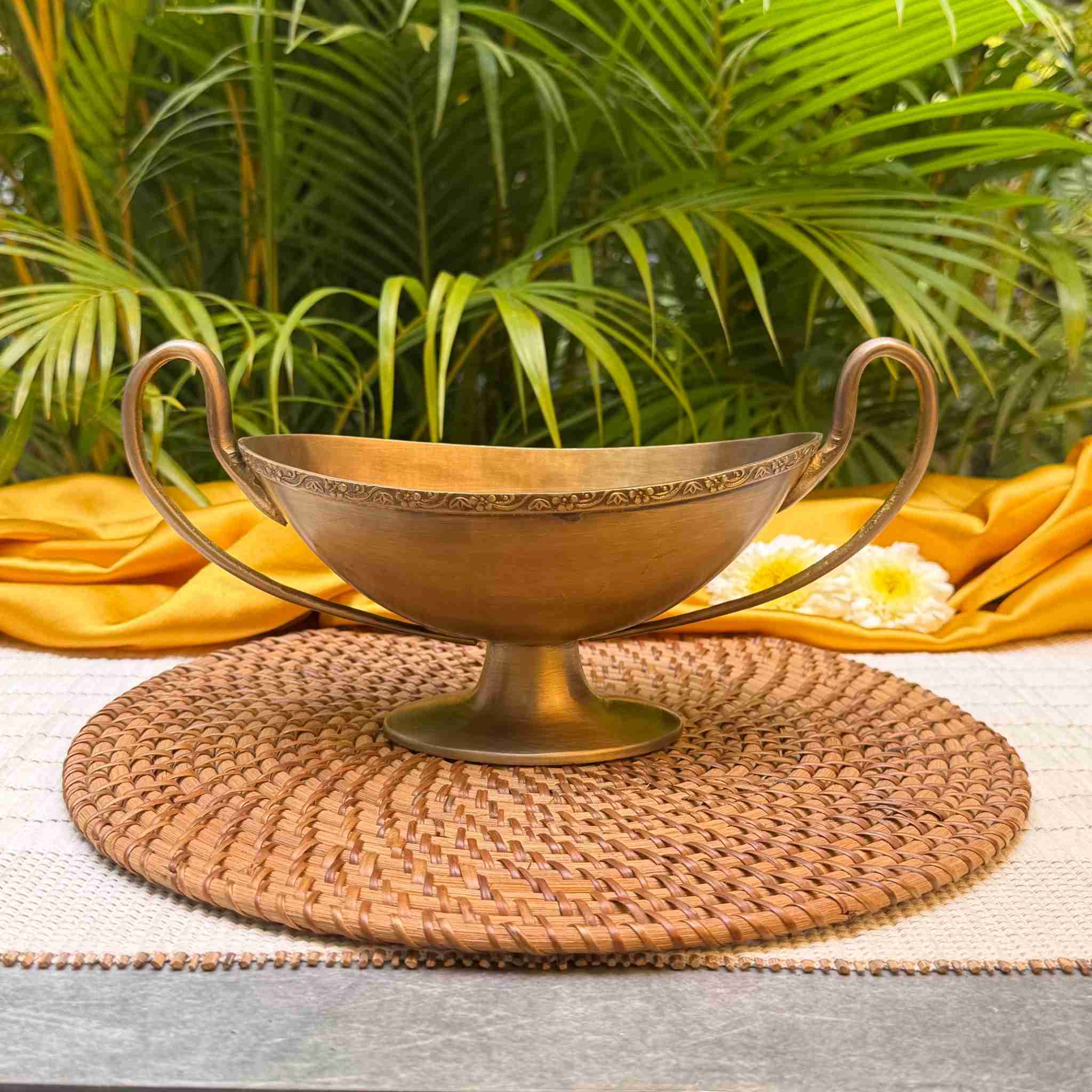 A brass pedestal bowl with two handles sits on a round woven placemat, styled with yellow fabric and white flowers against a backdrop of green palm leaves.