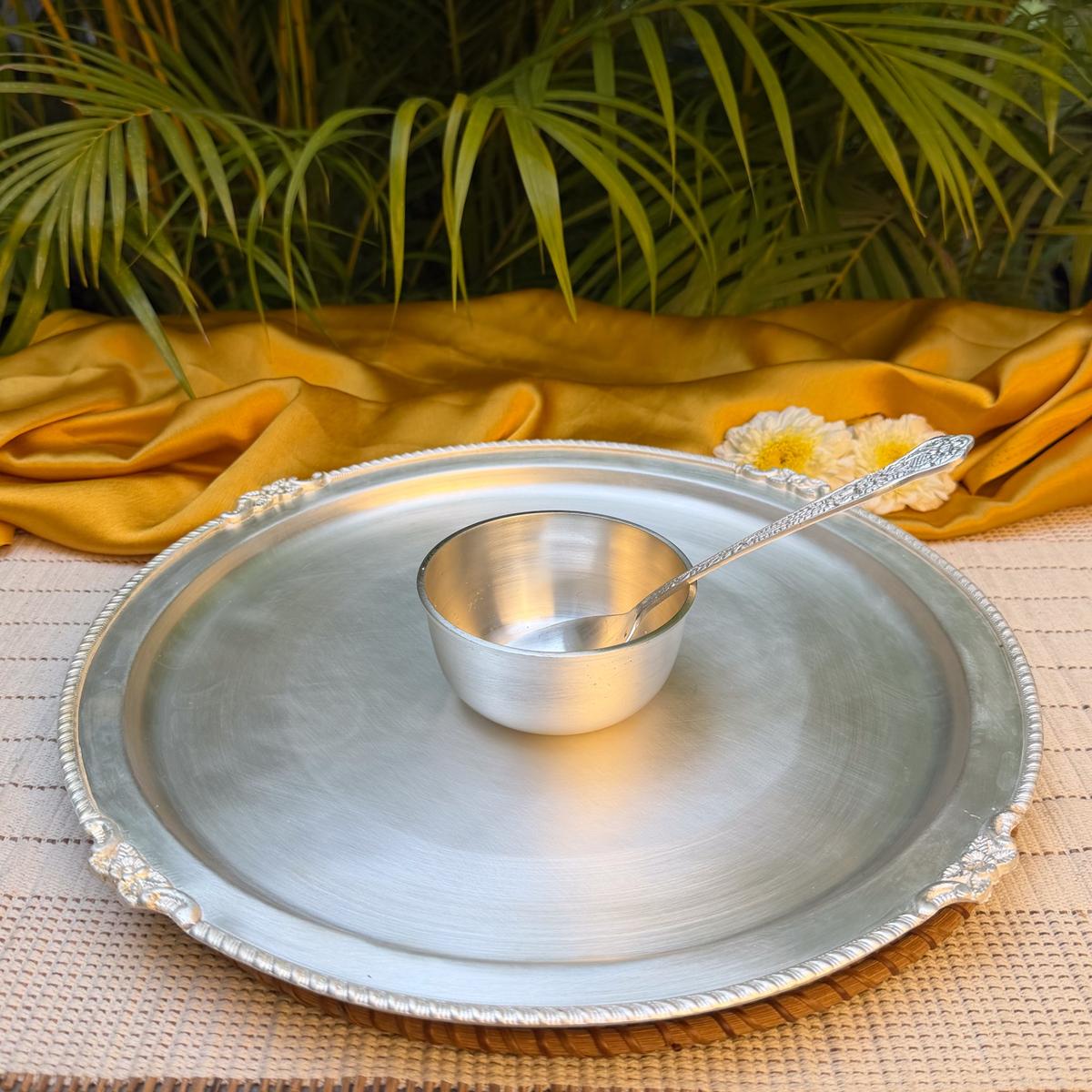 A silver-toned metal tray with ornate decorative edges holds a small matching metal bowl and spoon, styled on a woven mat with draped mustard-yellow fabric and green palm leaves in the background.